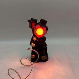 Könnte beinhalten: Vintage Keramiklampe in Form eines Mannes, der eine Straßenlaterne hält. Die Lampe ist dunkelbraun mit goldenen Akzenten und einem leuchtend gelben Licht im Gesicht. Die Lampe hat einen Zylinder und ein Kabel.