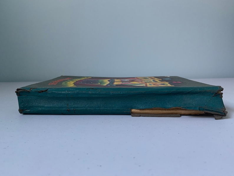 Puede incluir: Un libro antiguo de tapa dura verde con una ilustraci&oacute;n colorida en la portada. El libro est&aacute; ligeramente desgastado y tiene un desgarro en el borde inferior.