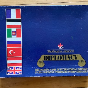Könnte beinhalten: Eine blaue Brettspielbox mit einem goldenen Schriftband, auf dem "DIPLOMACY" steht. Die Box zeigt ein kanadisches Flaggenlogo für Waddington Sanders und eine Reihe von Flaggen, die die verschiedenen Länder im Spiel repräsentieren.