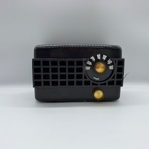 Könnte beinhalten: Ein schwarzes Vintage-Radio mit einem runden Zifferblatt und einem gitterartigen Lautsprecher. Das Zifferblatt hat Zahlen von 55 bis 160 und das Wort "TRUE" ist darauf gedruckt.