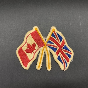 Op de afbeelding: Een vintage geborduurde patch met de gekruiste vlaggen van Canada en Groot-Brittannië. De vlaggen zijn op een witte achtergrond met rode en blauwe accenten.