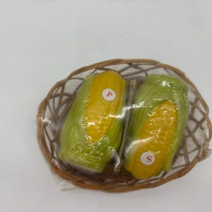 May include: A pair of corn-shaped salt and pepper shakers in a woven basket. The shakers are yellow and green, with the letters "P" and "S" on them. The basket is brown and oval-shaped.