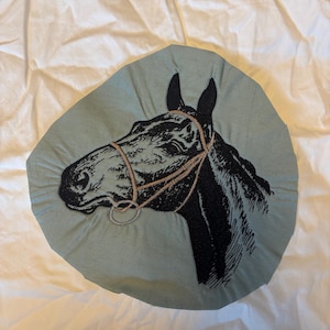 Könnte beinhalten: Ein hellblaues Zierkissen mit einem gestickten schwarzen Pferdekopf-Design. Der Pferdekopf ist mit Zaumzeug und Zügeln detailliert. Das Kissen befindet sich auf einem weißen Hintergrund.