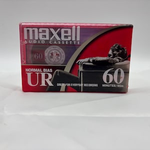 May include: A Maxell audio cassette tape in a red and black case. The case features the text "maxell AUDIO CASSETTE" and "60 MINUTES/90m". The cassette tape is labeled "NORMAL BIAS UR". The case has a graphic of a person.