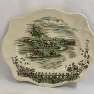 May include: A white oval serving platter with a green and brown country scene featuring a river, houses, and trees. The border of the platter is decorated with green vines and pink roses.