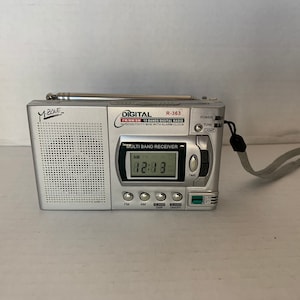 Puede incluir: Una radio digital AM/FM plateada con reloj despertador incorporado. La radio tiene una antena telescópica y una correa. La pantalla muestra "12:13" y "AM". La radio está etiquetada como "M.ZONE" y "DIGITAL FM/MW SW 6 Bands Digital Radio Sensitivity Mini with Alarm Clock".