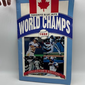 May include: A Toronto Blue Jays "World Champs" collector's edition featuring the Canadian flag and team photos. The cover has the text "Toronto Blue Jays" and "World Champs 1992". The bottom reads "Collector's Edition".