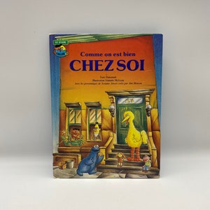 Puede incluir: Un libro de Plaza Sésamo titulado "Comme on est bien CHEZ SOI" con ilustraciones de Big Bird, Cookie Monster y otros personajes de Plaza Sésamo. La portada del libro presenta una casa amarilla con una puerta verde y un buzón rojo.