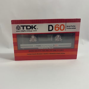 Puede incluir: Una cinta de cassette roja y negra con la etiqueta "TDK D60" con el texto "Normal Position Position normale" y "Low Noise High Output/Faible Bruit Fort Signal A La Sortie" impreso en la parte delantera. La cinta de cassette es un medio de grabación de audio vintage.