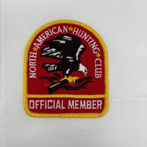 May include: Embroidered patch with a red background, featuring a black and white eagle design. The patch includes the text "NORTH AMERICAN HUNTING CLUB" and "OFFICIAL MEMBER" in yellow and white lettering, with a yellow border.