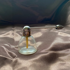 May include: A clear glass atomizer with a silver top and a long silver nozzle. The atomizer is sitting on a brown fabric surface.