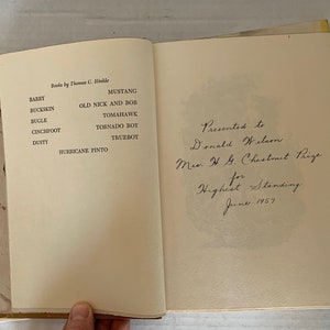 May include: A book titled "Books by Thomas C. Hinkle" with a list of horse names, including Barry, Buckskin, Bugle, Cinchfoot, Dusty, Hurricane Pinto, Mustang, Old Nick and Bob, Tomahawk, Tornado Boy, and Trueboy. The book is inscribed "Presented to Donald Wilson Ms. H. G. Chestnut Prize for Highest Standing June 1957."