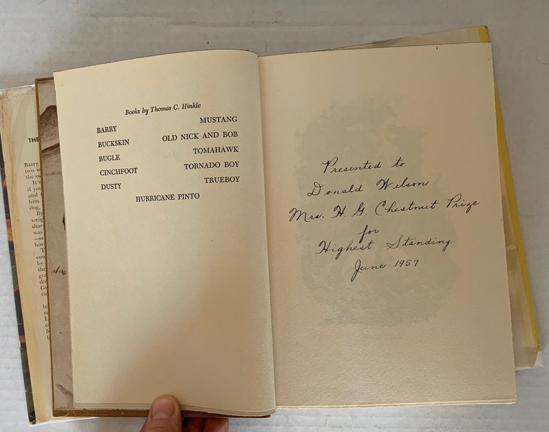 May include: A book titled "Books by Thomas C. Hinkle" with a list of horse names, including Barry, Buckskin, Bugle, Cinchfoot, Dusty, Hurricane Pinto, Mustang, Old Nick and Bob, Tomahawk, Tornado Boy, and Trueboy. The book is inscribed "Presented to Donald Wilson Ms. H. G. Chestnut Prize for Highest Standing June 1957."