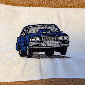 Peut inclure: Image brodée d'une voiture classique bleue avec une calandre et des roues noires. La voiture est représentée sur un fond blanc. La broderie détaille les caractéristiques de la voiture, y compris la prise d'air et les phares.