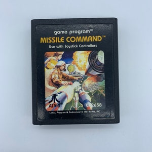 May include: Black Atari Missile Command game cartridge. The label features a pilot in a cockpit, missiles, and a city under attack. Text includes "MISSILE COMMAND" and "Use with Joystick Controllers."