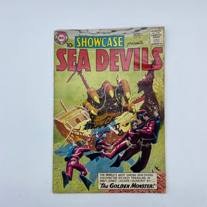 Puede incluir: Una portada de cómic vintage con una ilustración colorida de un barco hundido con buzos y una criatura marina monstruosa. El texto de la portada dice "Showcase presenta Sea Devils" y "The Golden Monster!"