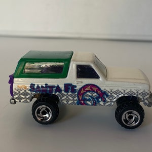 May include: White toy truck with a green roof and "SANTA FE" printed on the side. The truck has a black and white geometric pattern and a purple and pink design.