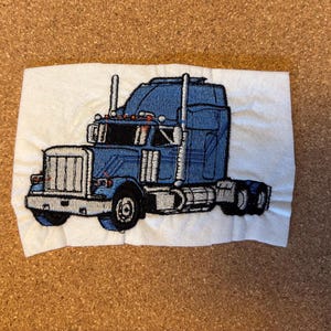 May include: Embroidered patch showing a blue articulated lorry with white and black detailing. The lorry has a detailed design, including the cab, trailer, and wheels. The patch is on a white fabric background.