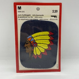 May include: An iron-on patch featuring a colourful embroidered image of a Native American wearing a headdress. The patch is on a blue background and is packaged in a clear plastic bag with a red and white label. The label includes the text "zum Aufbügeln 100% Baumwolle à appliquer au fer 100% coton da applicare con il ferro 100% cotone" and the price "2.20".