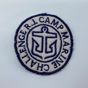 Può includere: Una toppa ricamata bianca con un'ancora blu e il testo "Challenge R.L. Camp Marine" attorno al bordo.