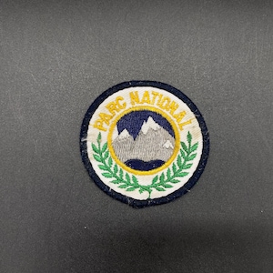 May include: A white, round embroidered patch with a blue circle in the center. The circle has a yellow border and depicts a white mountain range with a blue lake. The text "PARC NATIONAL" is embroidered in yellow around the circle.