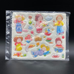 May include: A sheet of colourful stickers featuring various characters, including a girl wearing a red dress, a girl wearing a blue dress, a girl wearing a yellow dress, and a girl wearing a green dress. The stickers are ideal for adding a touch of whimsy to any project.