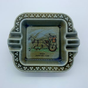 May include: A green and white ceramic ashtray with a shamrock border. The center of the ashtray features a colorful illustration of a horse-drawn jaunting car with the text "IRISH JAUNTING-CAR".