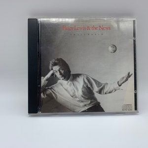 Op de afbeelding: Een cd-hoes met een zwarte rug en een witte achtergrond. De voorkant toont een zwart-witfoto van Huey Lewis, de zanger van Huey Lewis and the News, met de bandnaam en de albumtitel "Small World" in rood.