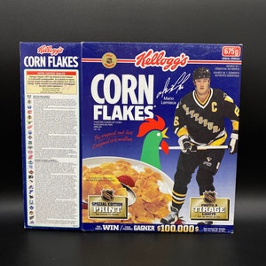 Könnte beinhalten: Eine blaue und weiße Kellogg's Corn Flakes-Müslipackung mit einem Hockeyspieler, der ein Trikot der Pittsburgh Penguins trägt. Die Packung hat einen Special Edition Print des NHL-Logos und einen Wettbewerb, bei dem man 100.000 $ gewinnen kann.