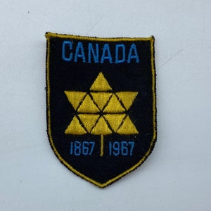 Puede incluir: Un parche de fieltro negro con una hoja de arce estilizada amarilla y el texto "CANADA 1867 1967" en azul.