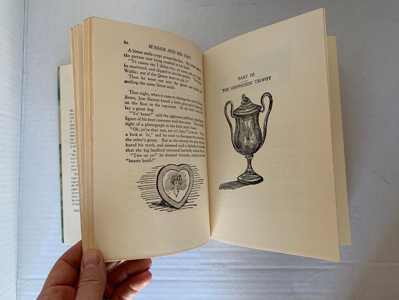 May include: A book open to a page with text about a dog and a trophy. The text reads "Madam and His Coat" and "The Shepherd's Trophy". There is a black and white illustration of a dog and a trophy.