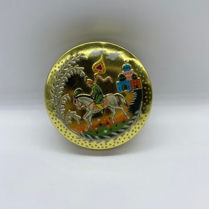 May include: A round, gold-coloured tin featuring a white horse and rider in a colourful scene. The scene includes a building with a dome and a heart-shaped object in the sky.