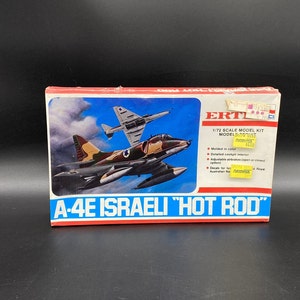 Puede incluir: Una caja de kit de maqueta para un avión de combate A-4E israelí "Hot Rod". La caja presenta una ilustración colorida del avión en vuelo. El texto en la caja dice "ERTL 1/72 Scale Model Kit Models of Merit 1650".