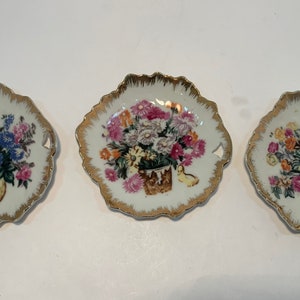 May include: Set of three small decorative plates with gold trim and floral designs. Each plate features a different bouquet of flowers in a vase.