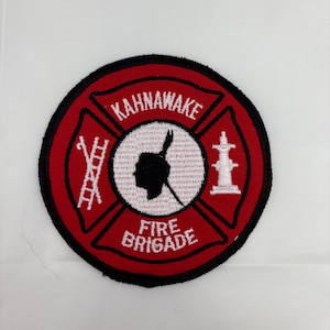 Könnte beinhalten: Ein runder Aufnäher mit rotem Rand und weißer Mitte. Der Aufnäher zeigt oben den Text "KAHNAWAKE" und unten "FIRE BRIGADE". Im Inneren des Aufnähers befinden sich eine Silhouette, eine Leiter und ein Hydrant.