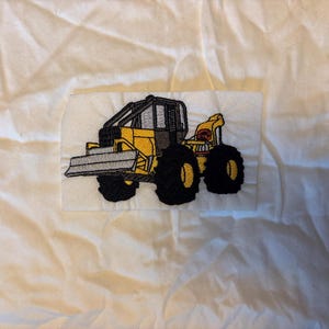 May include: Embroidered patch featuring a yellow and black construction vehicle with a blade and large tires. The patch is rectangular and sewn onto a white fabric background. The vehicle has a detailed design.