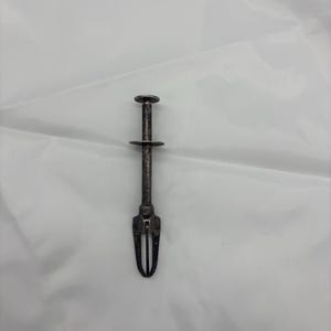 Może przedstawiać: Zabytkowy, srebrzysty serwer do ciasta. Posiada uchwyt, okrągły środek i dwuzębowy widelec na dole. Serwer jest przeznaczony do podnoszenia i serwowania ciast lub małych ciast. Tło to biała, pomarszczona tkanina.