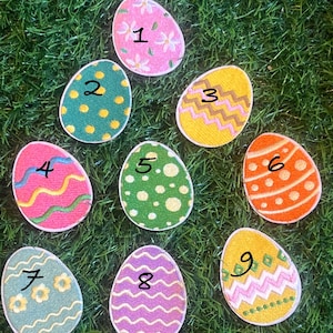 May include: Nine embroidered felt Easter eggs with different patterns and numbers 1 through 9. The eggs are on a green grass background.
