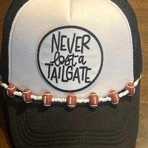 May include: Black and white trucker hat with a white front panel. The front panel features a circular patch with the text "NEVER lost a TAILGATE". A beaded chain of football-shaped beads adorns the brim.