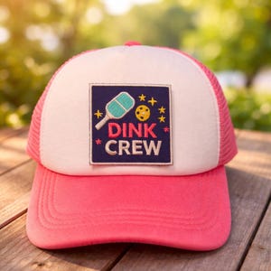 May include: A pink and white trucker hat with a square patch reading "DINK CREW." The patch features a pickleball paddle and ball design. The hat has a pink peak and mesh back.