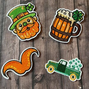 May include: Four embroidered patches with a St. Patrick's Day theme. Includes a leprechaun face, a beer mug with foam and a shamrock, an orange swirl, and a green truck filled with shamrocks. All patches have white borders.