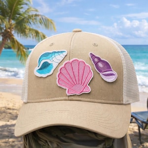 Puede incluir: Gorra de camionero beige con la parte trasera de malla blanca y tres parches de conchas marinas en azul claro, rosa y morado. La gorra se presenta sobre un fondo de playa.