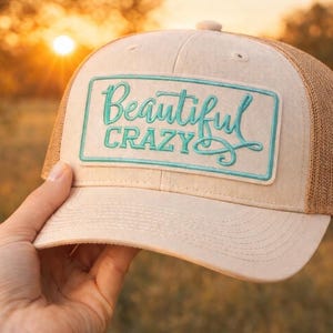 May include: A beige trucker hat with a mesh back and a curved brim. The hat features a rectangular patch with the words "Beautiful Crazy" in teal script. The hat is held up against a blurred background.