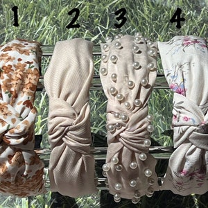 May include: Four different styles of knotted headbands. The first headband is white with a brown floral pattern. The second headband is a solid beige color. The third headband is beige with white pearl embellishments. The fourth headband is white with a light pink floral pattern.