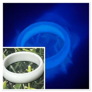 May include: A white silicone ring that glows blue under a blacklight. The ring is shown on a green grassy surface.