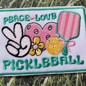 Könnte beinhalten: Gestickter Aufnäher mit den Worten "PEACE LOVE PICKLEBALL". Der Aufnäher zeigt ein Friedenszeichen, ein rosa Herz, eine gelbe Blume, einen Pickleball-Schläger und einen gelben Pickleball. Der Rand ist grün.