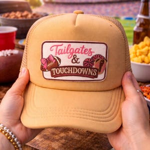 May include: Tan trucker hat with a mesh back and a patch that reads "Tailgates & Touchdowns." The patch features a football, a bow, and a football-shaped donut. The hat is held up by a person.