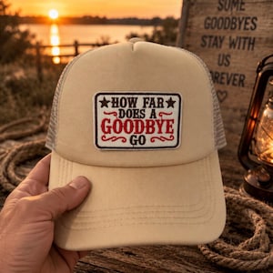 May include: A beige trucker hat featuring a rectangular patch on the front. The patch has a white background with a brown border and the text "HOW FAR DOES A GOODBYE GO" in brown and red lettering, accented with two small stars and decorative swirls.