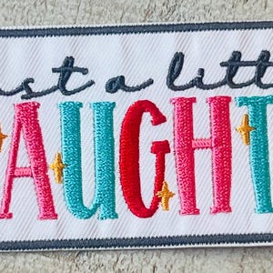 May include: A white embroidered patch with gray stitching around the edges. The patch says "Just a little naughty" in colorful letters with gold glitter accents.