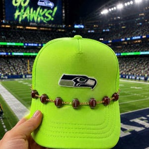 May include: A neon green baseball cap featuring a Seattle Seahawks logo patch on the front crown. A gold-toned chain is wrapped around the base of the crown, decorated with small brown football-shaped beads with white stitching details.